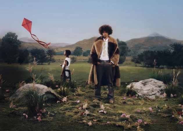 Man in fur coat standing in a grassy field with mountains in the background and a person flying a kite