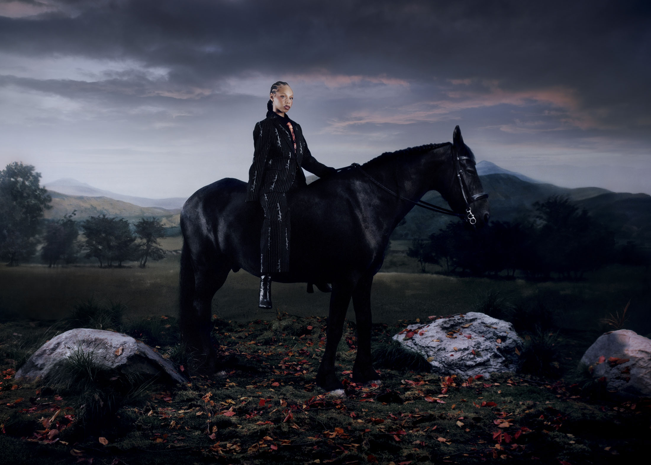 Model wearing a dark pinstripe suit mounted on a black horse in a dramatic landscape under an overcast sky.