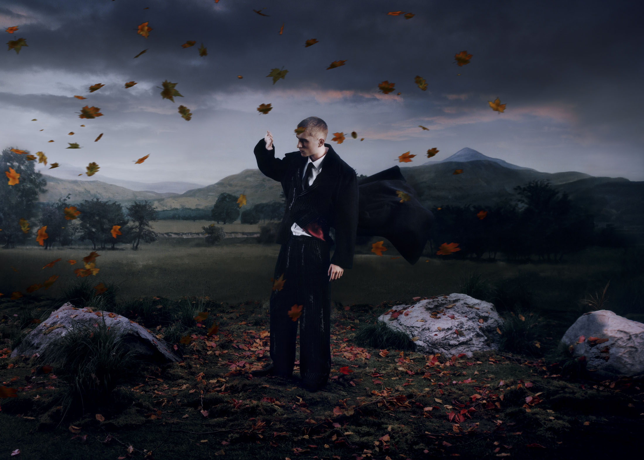 Man in dark suit standing in a dramatic rural landscape surrounded by falling autumn leaves captured by Elliot Hensford
