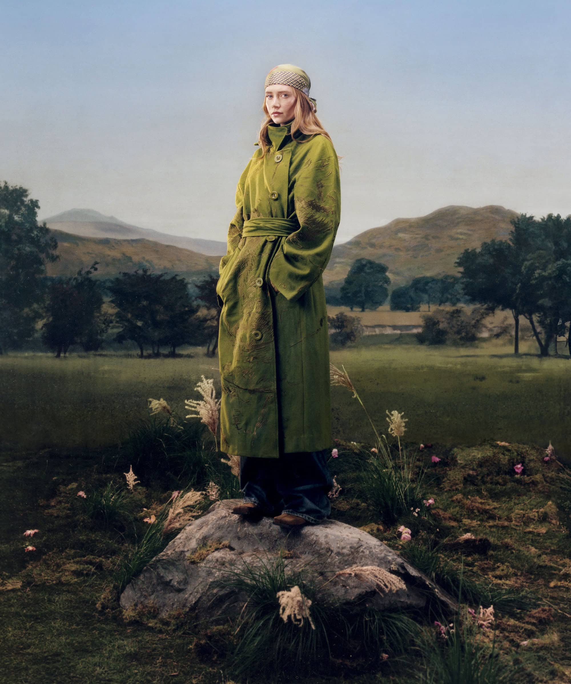 Model standing on a mossy rock wearing an olive green belted trench coat against a misty valley landscape.