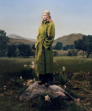 Model standing on a mossy rock wearing an olive green belted trench coat against a misty valley landscape.