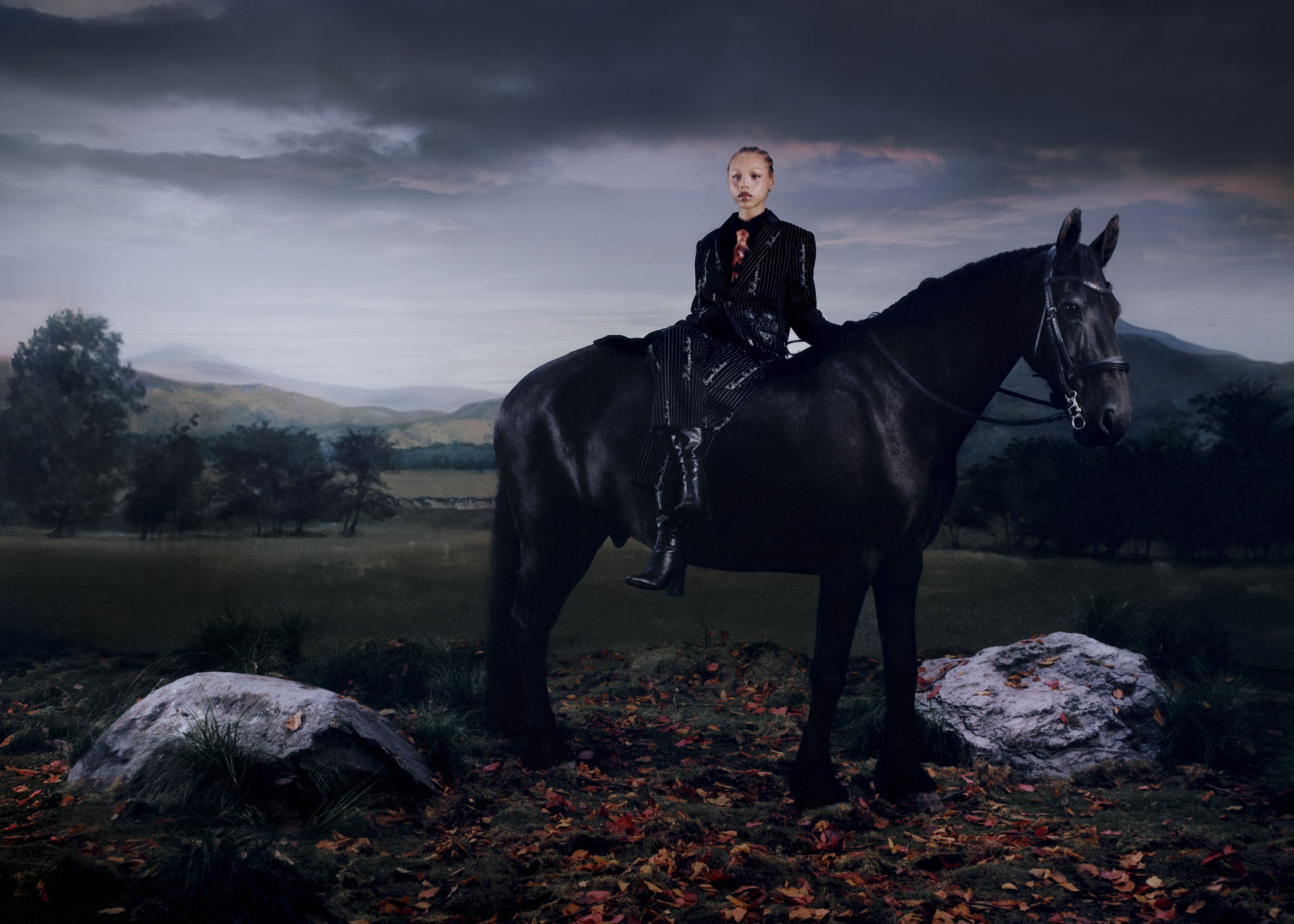 Model seated on a black horse in a landscape setting with rolling hills and a cloudy sky for KidSuper Fall 2025 campaign