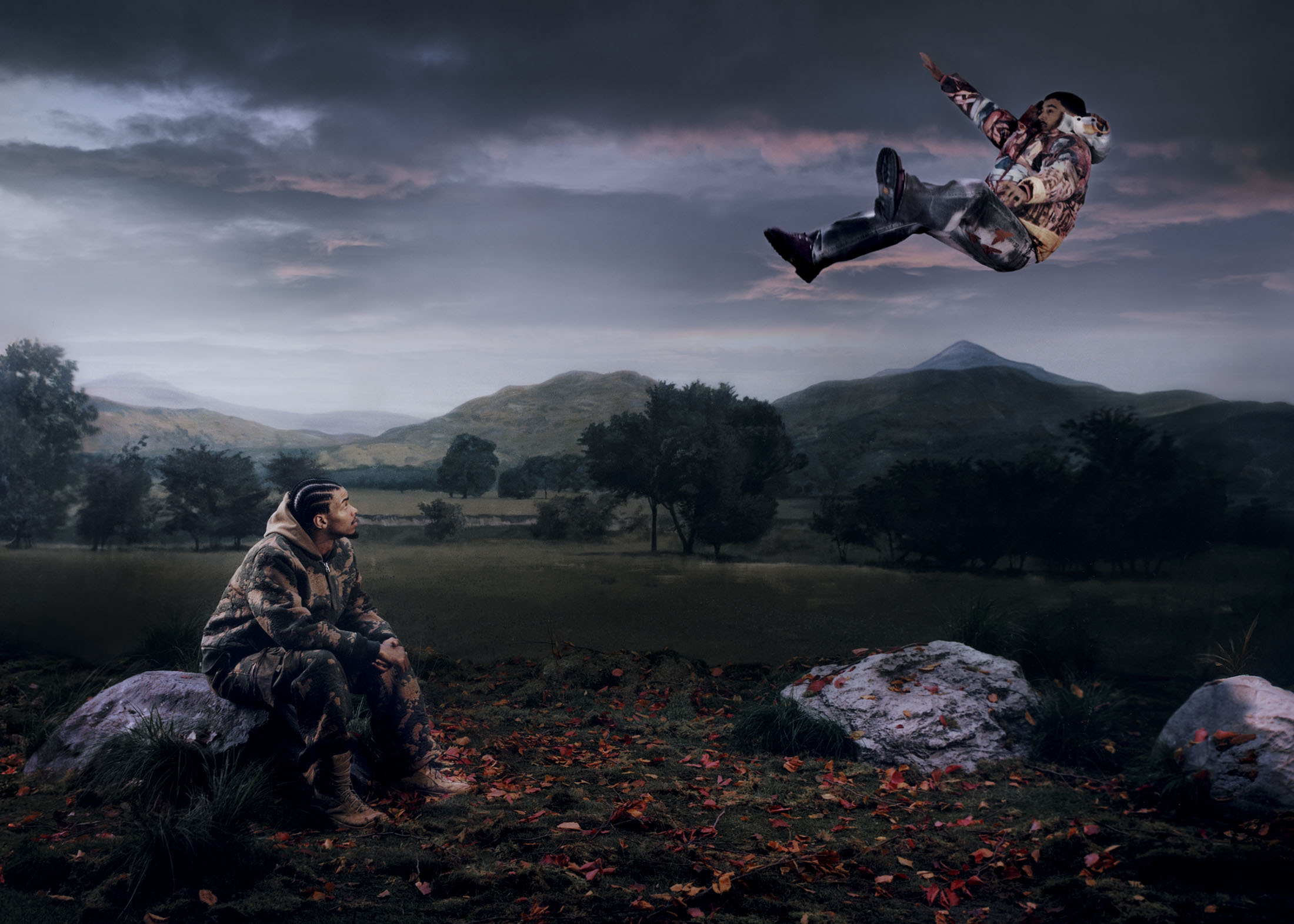 Man seated on a rock looking up at another man floating horizontally in the air against a moody landscape under a dramatic cloudy sky.