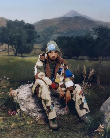 Model wearing a patterned puffer jacket and beanie seated on a rock in a landscape with hills and trees