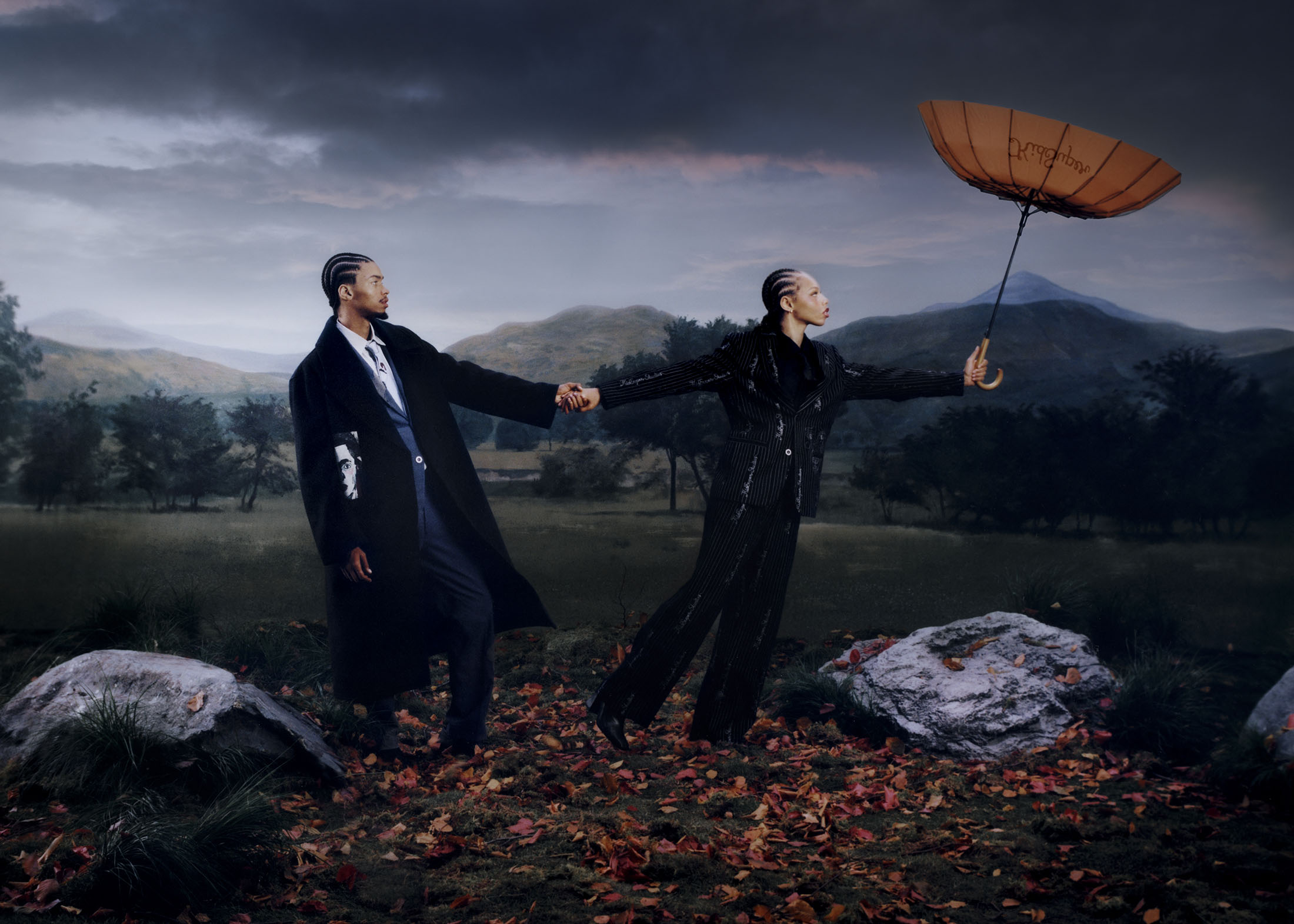 Two models standing in a moody landscape holding hands under an umbrella for KidSuper Fall 2025 campaign