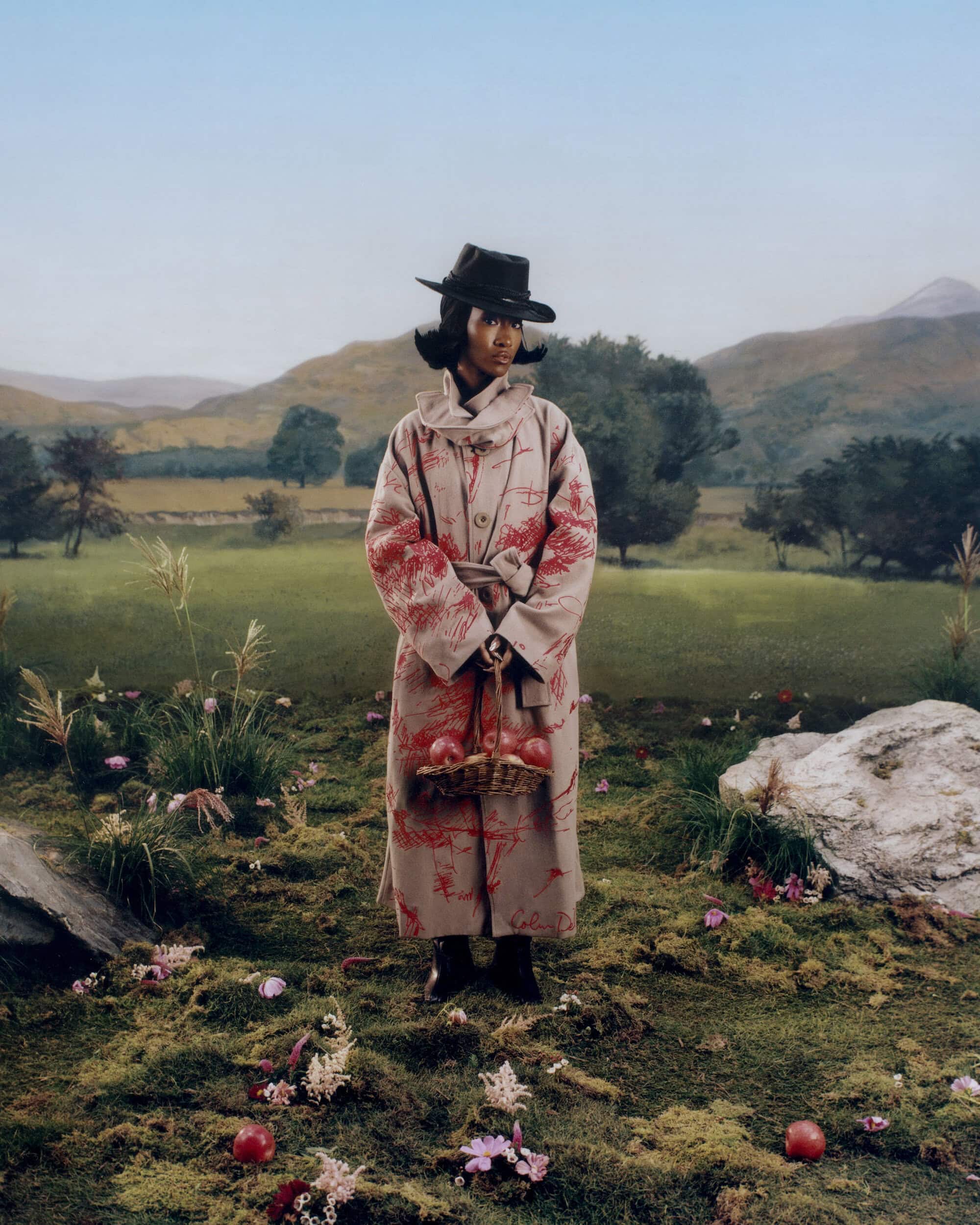 Model wearing printed long coat and black hat holding a wicker basket standing in a green landscape