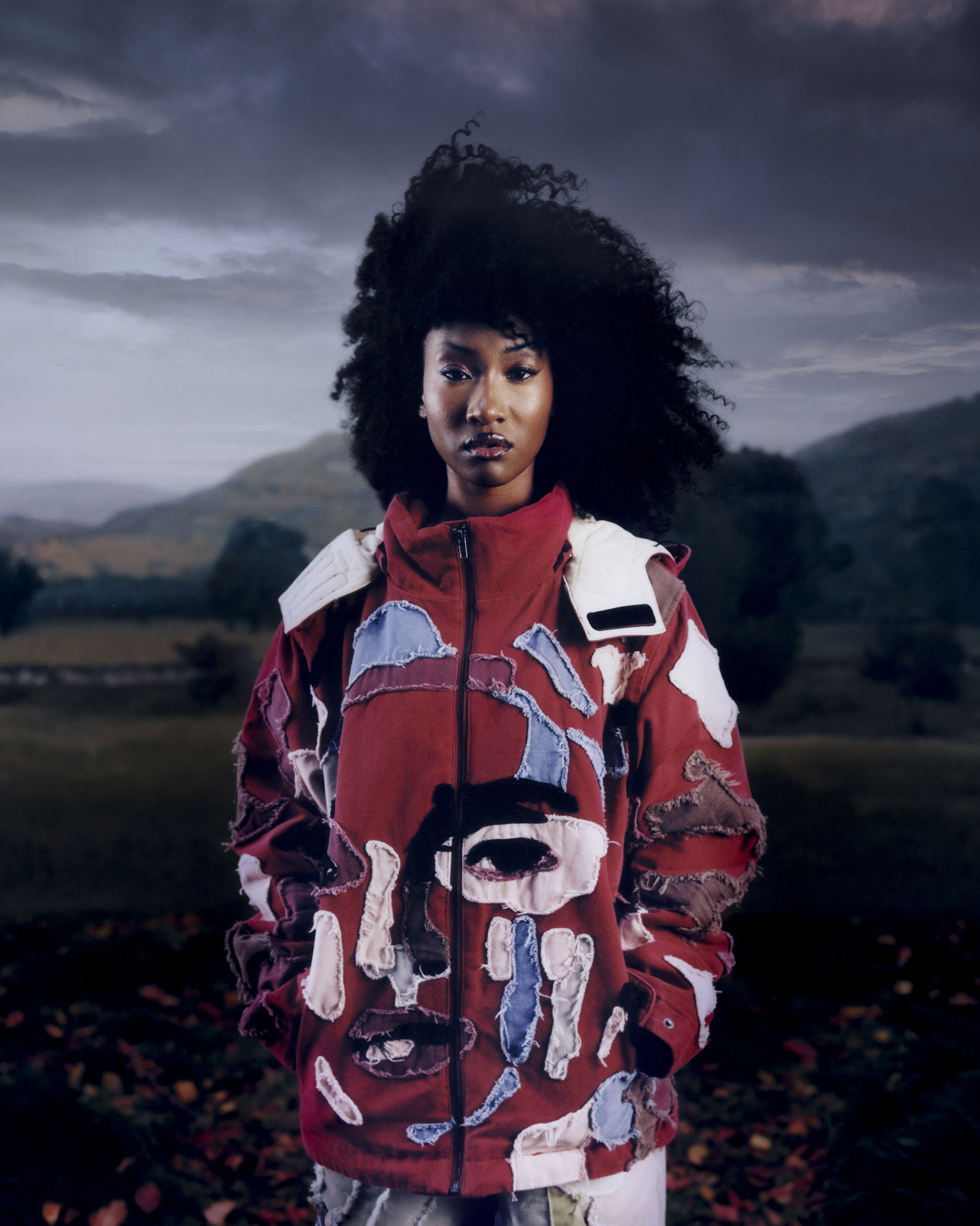 Model with voluminous dark curly hair wearing a patchwork red jacket outdoors against a mountainous landscape for KidSuper.