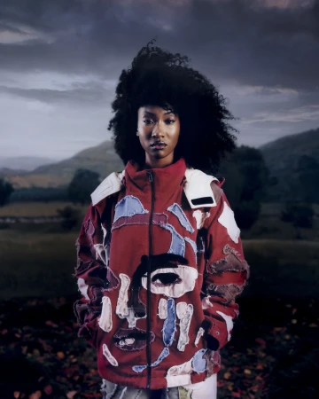 Model with voluminous dark curly hair wearing a patchwork red jacket outdoors against a mountainous landscape for KidSuper.