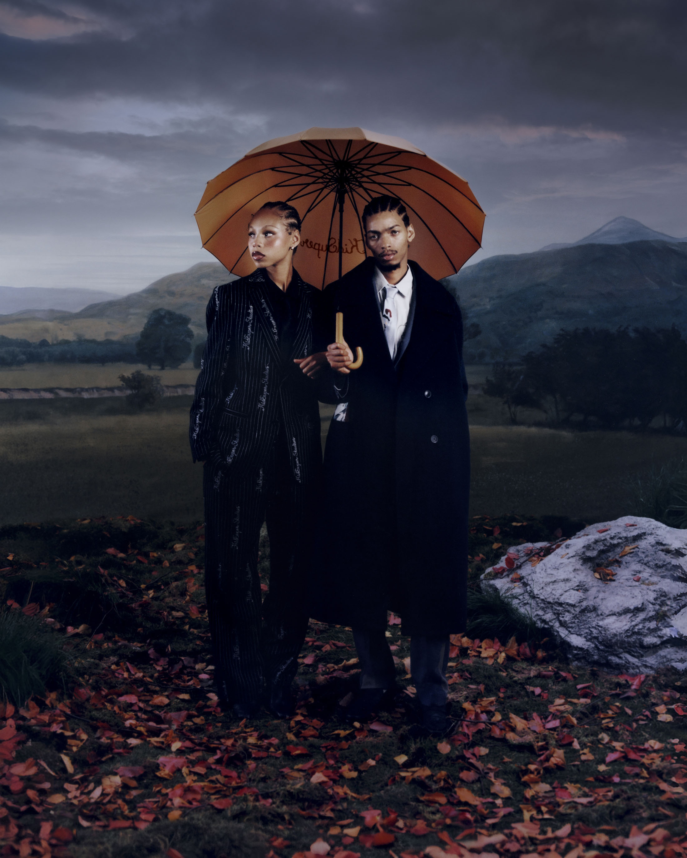 Two models standing under a large umbrella over a field of autumn leaves against a landscape background with hills