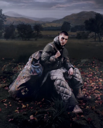 Man in padded quilted coat and trousers seated outdoors in a rural landscape at dusk