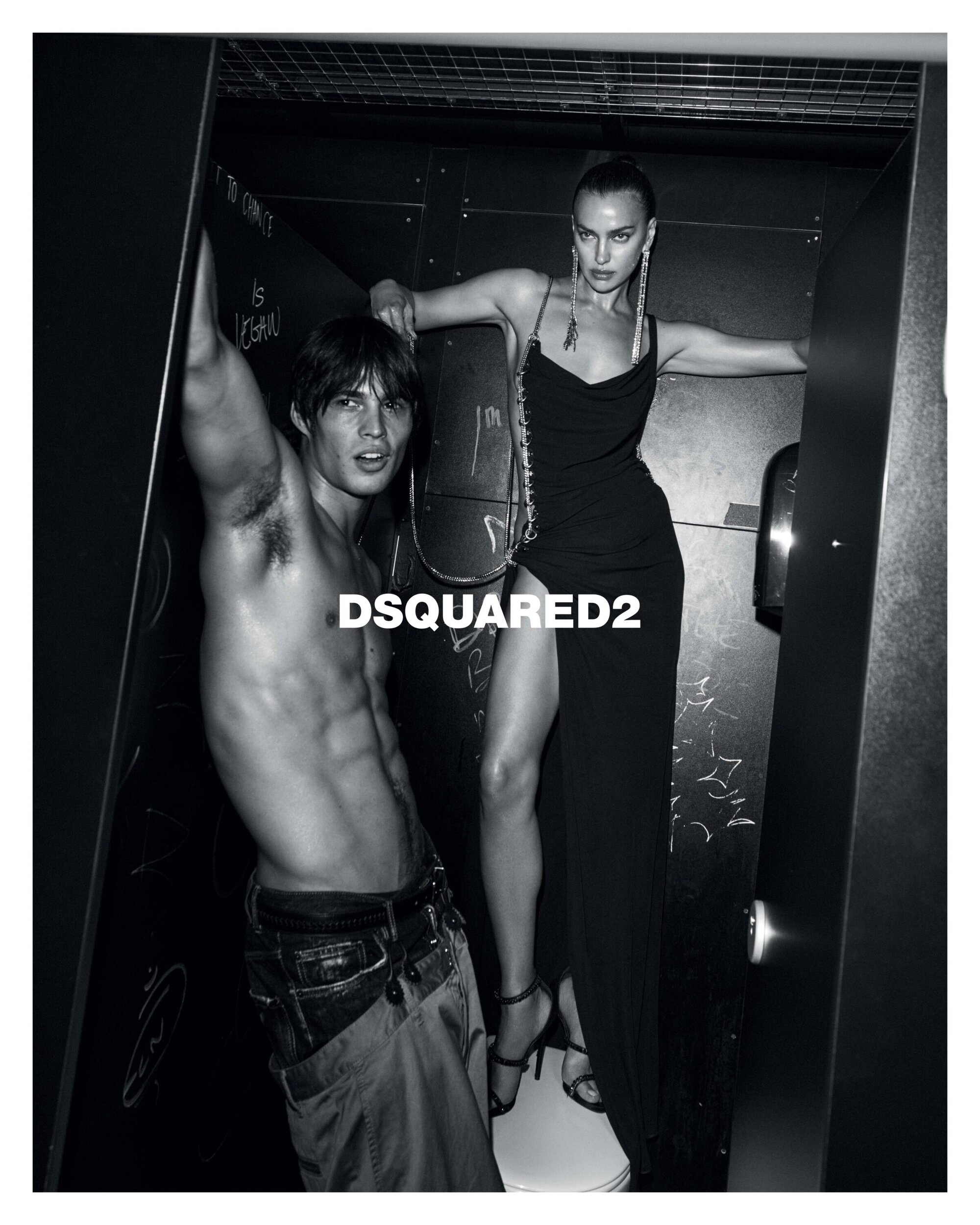 Black and white ad campaign image featuring Irina Shayk and a male model posed within a confined interior setting