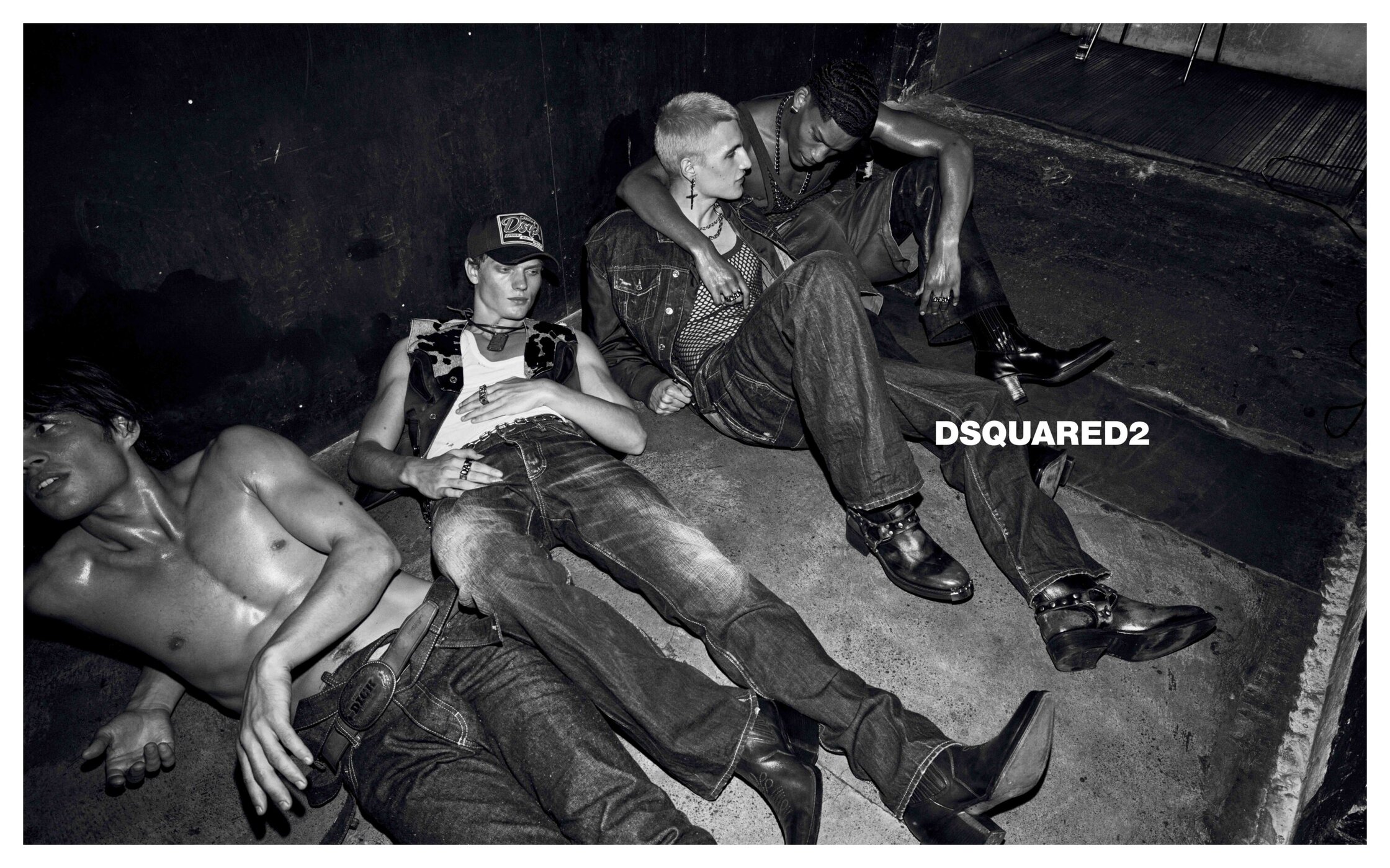 Monochrome campaign image of four young male models lounging on a dark concrete floor wearing denim and leather boots for Dsquared2.
