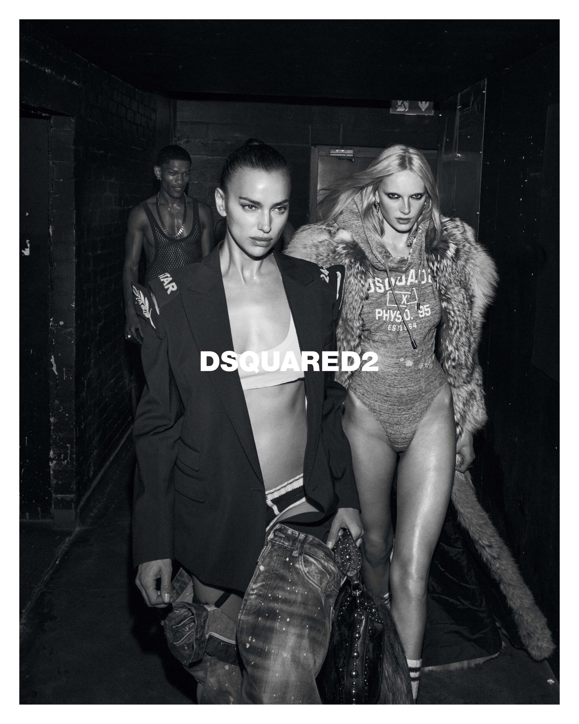 Black and white fashion campaign image featuring models Irina Shayk and Alex Consani walking in a dark corridor