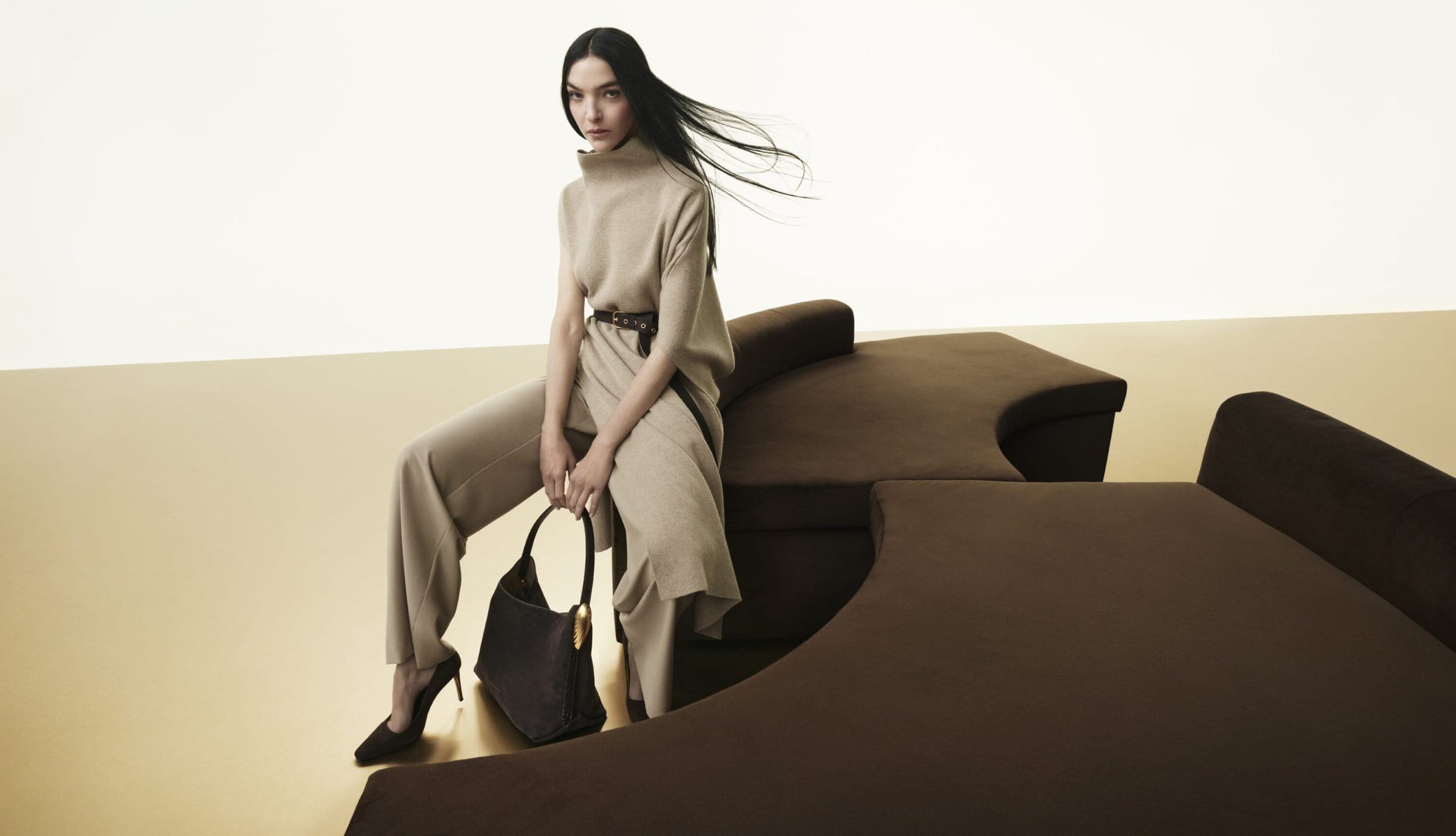 Woman in neutral knitwear sitting on dark brown sectional sofa holding a dark handbag against a minimal beige and white backdrop