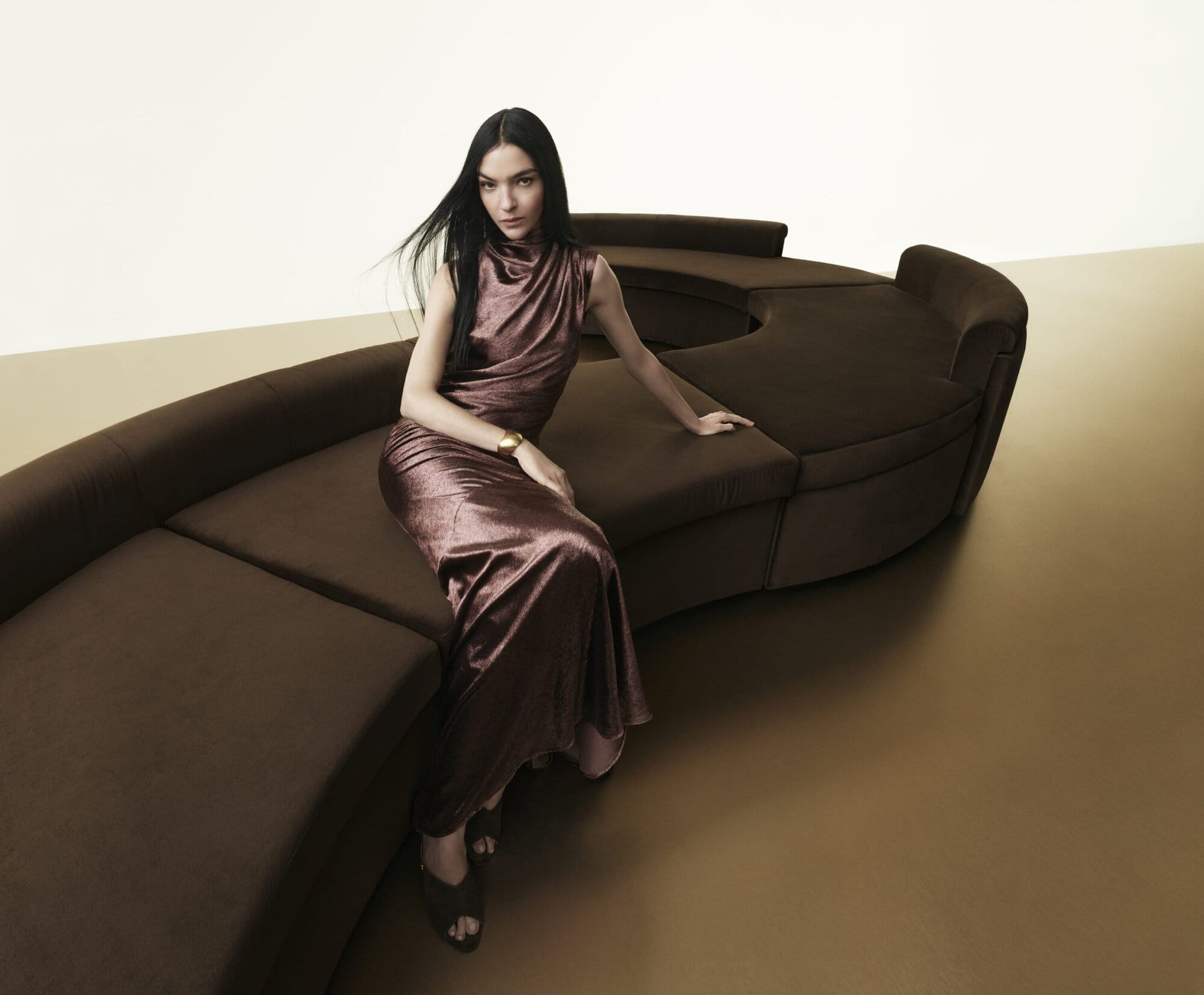 Model seated on a curved brown sofa wearing a metallic brown sleeveless gown and gold cuff bracelet