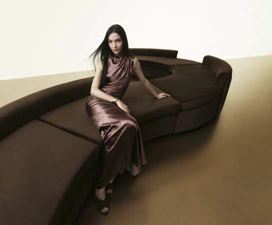 Model seated on a curved brown sofa wearing a metallic brown sleeveless gown and gold cuff bracelet