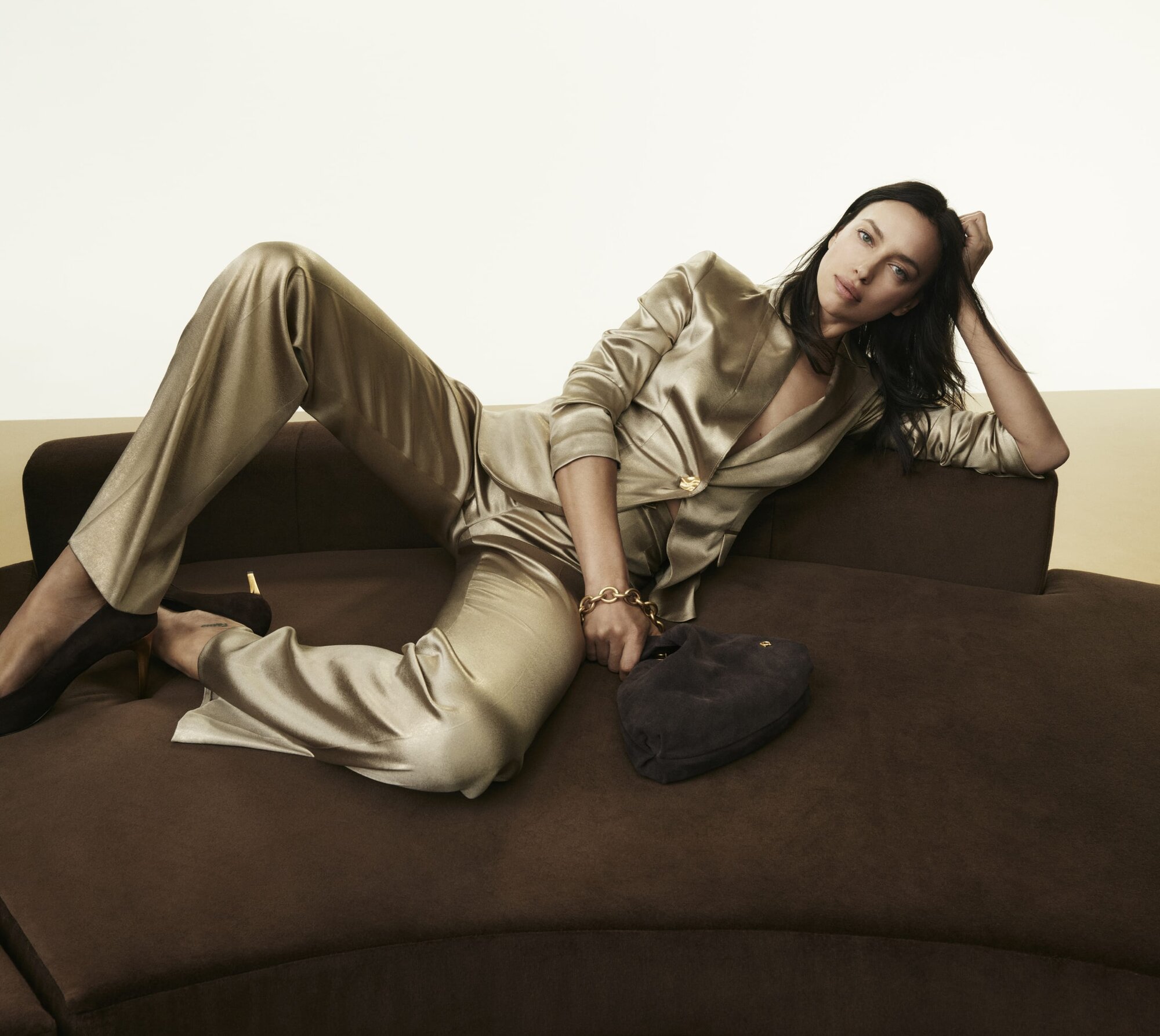 Model Irina Shayk reclining on a dark brown curved sofa wearing a gold satin pantsuit and holding a dark handbag.