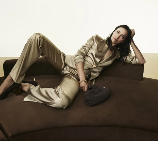 Model Irina Shayk reclining on a dark brown curved sofa wearing a gold satin pantsuit and holding a dark handbag.