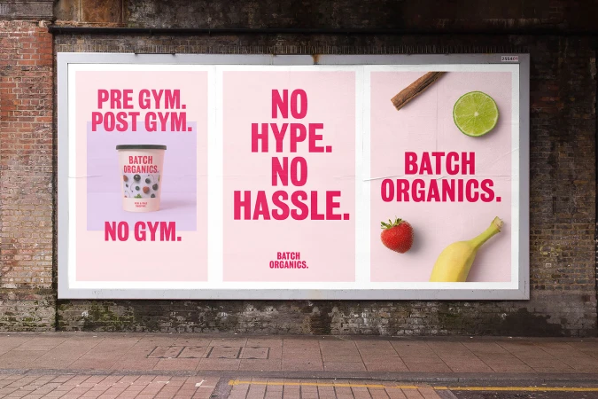 Billboard displaying three pink advertising posters for Batch Organics mounted on a weathered brick wall above a paved street.