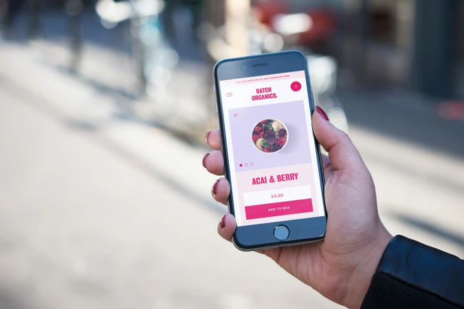 Hand holding a smartphone displaying the Batch Organics mobile app interface outdoors with a blurred street background.
