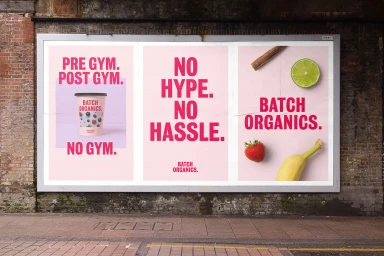 Billboard displaying three pink advertising posters for Batch Organics mounted on a weathered brick wall above a paved street.