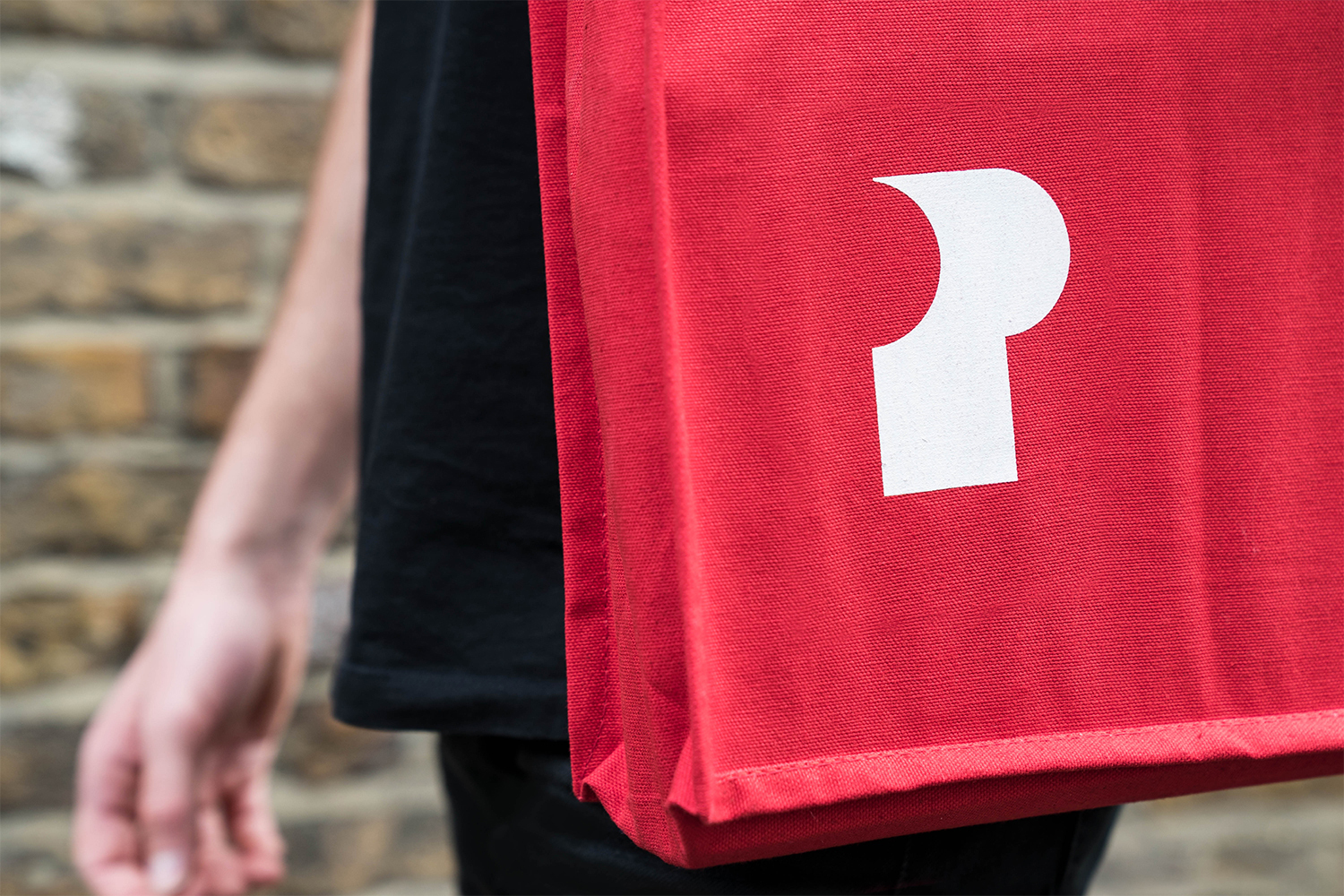 Close-up of a person wearing a bright red canvas tote bag with a white graphic logo against a blurred brick wall background.