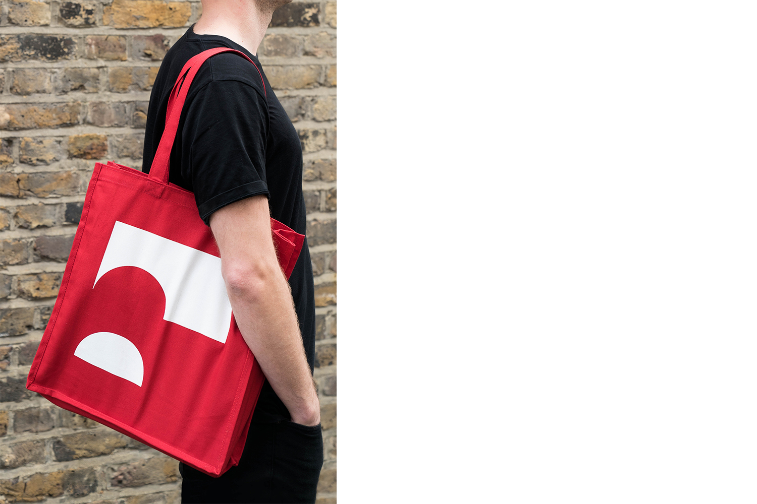 A person wearing a black t-shirt carries a bright red tote bag with a white graphic design against a brick wall.