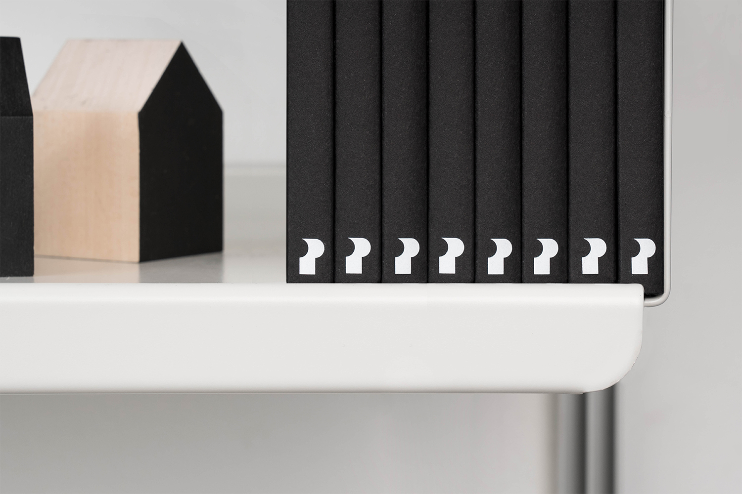 Close-up of black books with white typographic symbols on spines lined up on a white shelf next to wooden house models.