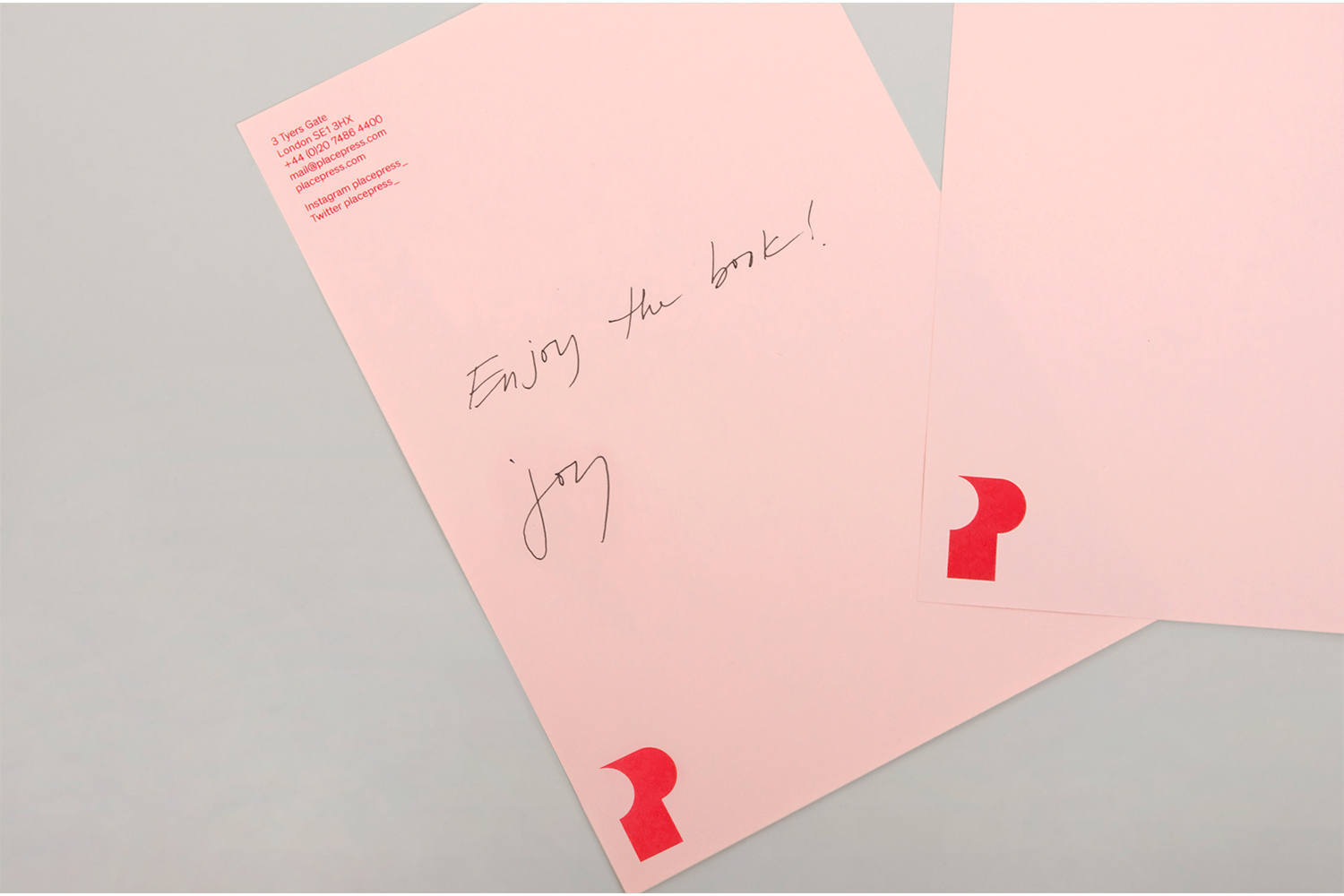 Two pink stationery sheets with red letterhead logos and handwritten black ink message resting on a neutral grey surface.