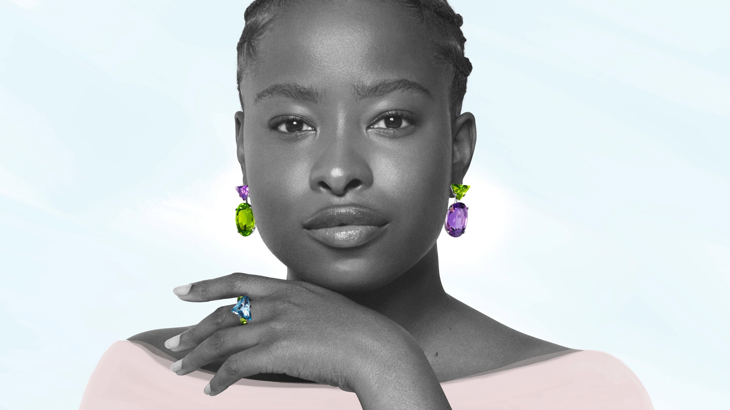 Close-up portrait of a Black woman wearing statement gemstone earrings and a ring, set against a light background.