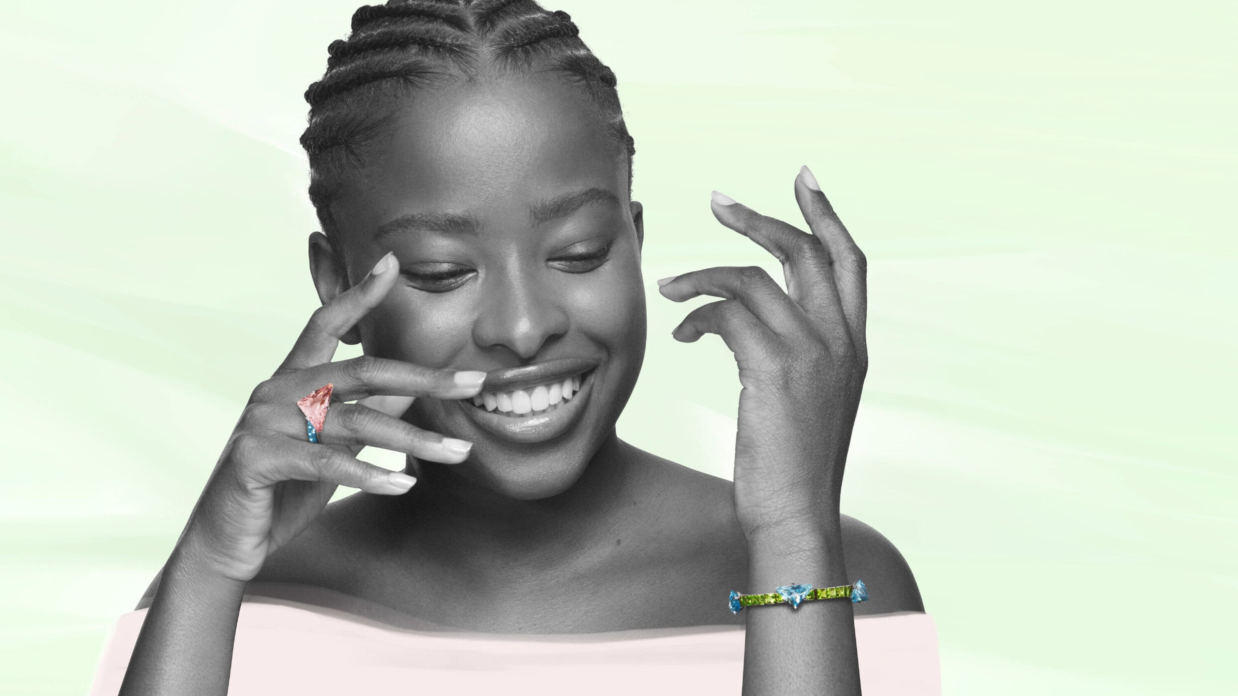 Black woman with braided hair smiling, wearing a colorful crystal ring and bracelet against a pale green and pink studio background.