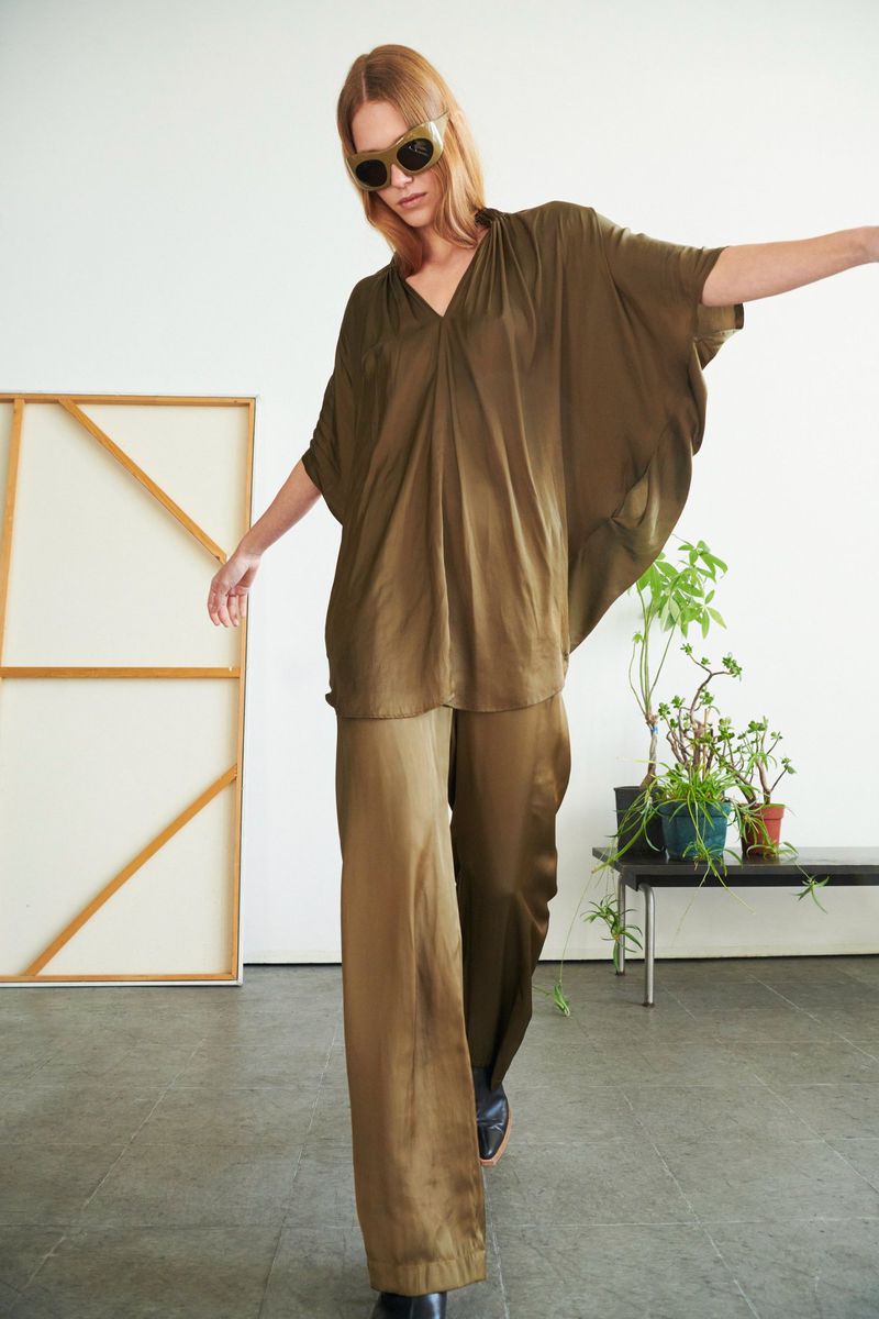 A woman in a flowing olive-green ensemble and vintage sunglasses poses in front of a canvas and plants in a light-filled studio.