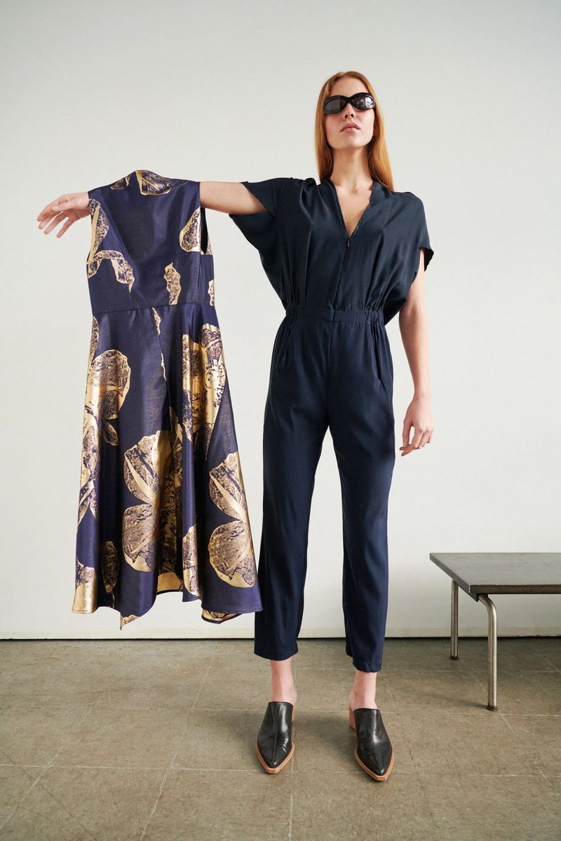Adult woman in dark jumpsuit and sunglasses poses with dress indoors in a fashion shot. Mules and a simple table are also present.