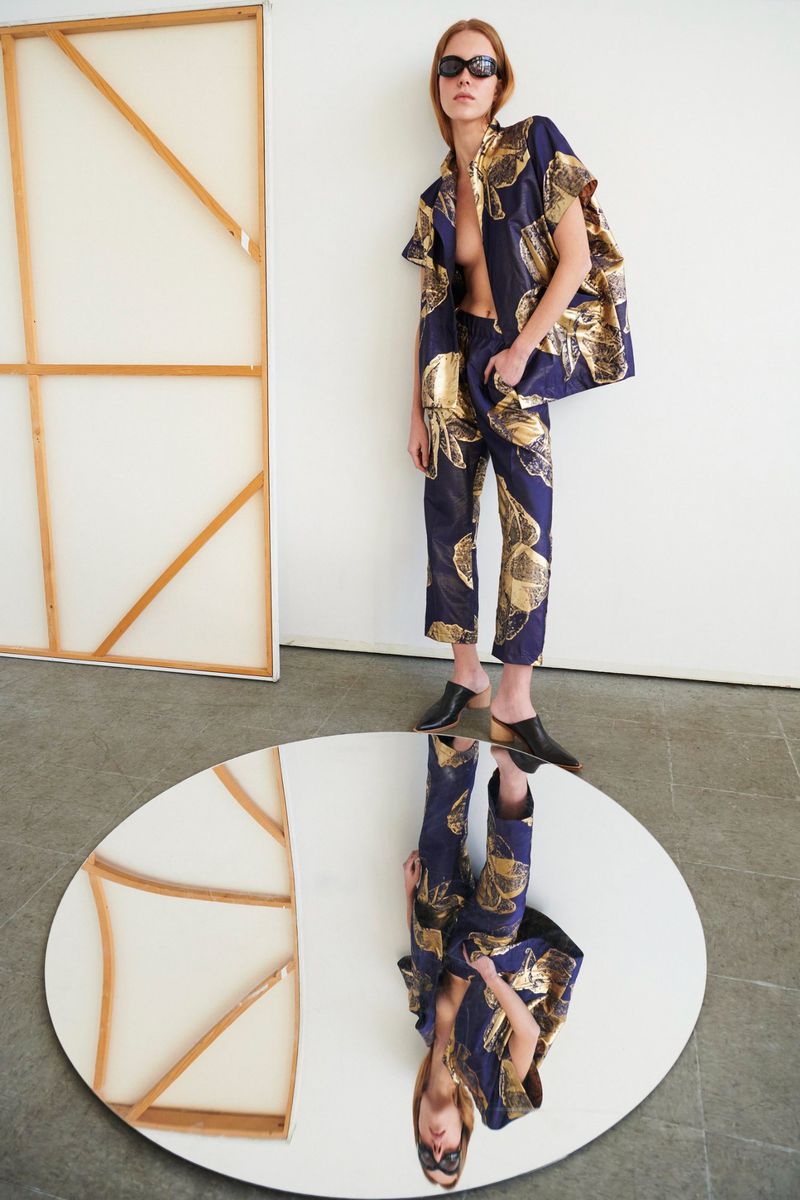 Fashion shot of an adult woman posing in a navy and gold brocade suit with black mules next to an oval mirror and artwork.