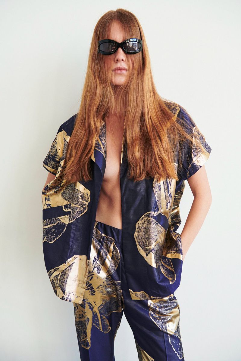 Stylish woman with long red hair wearing sunglasses and a blue and gold patterned two-piece outfit against a neutral background.