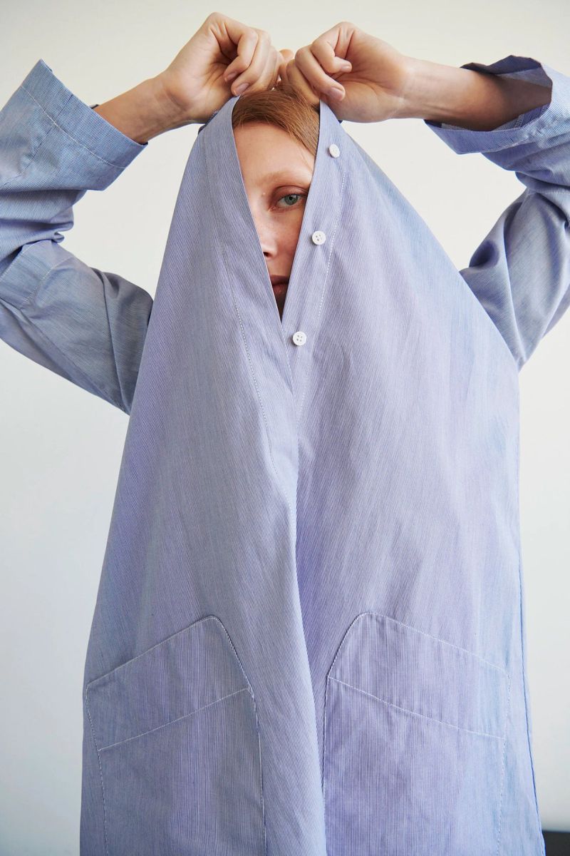 Studio shot of a redhead woman partially covered by a light blue button-down shirt, looking directly at the camera. Fashion portrait.