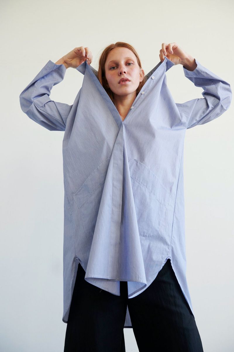 An adult woman poses in a photo studio, wearing a blue and white striped oversized shirt dress and black pants against a white backdrop.