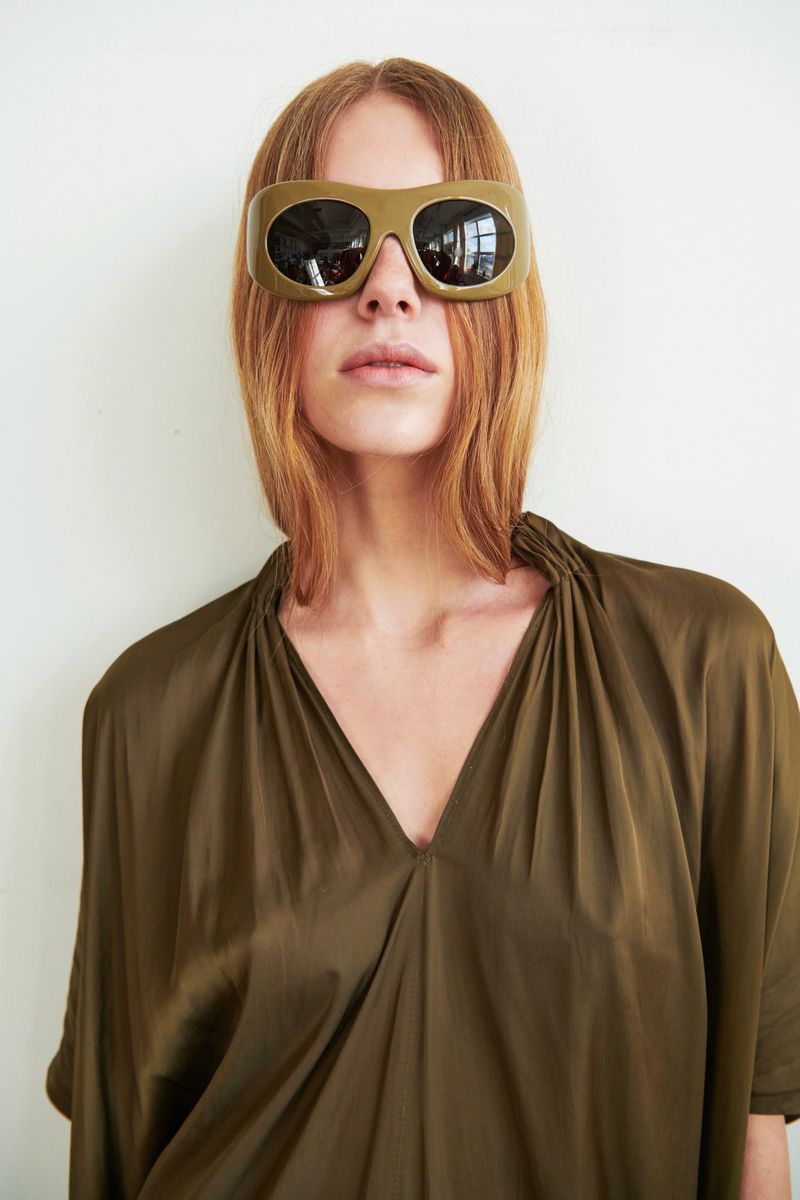 Woman with shoulder-length auburn hair wears oversized sunglasses and an olive-green dress against a plain white wall in soft lighting.