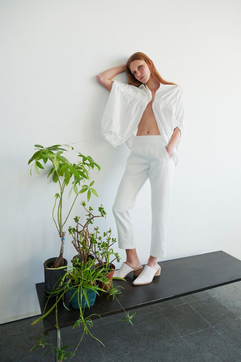 A woman in a white blouse and pants poses indoors next to potted plants, with a white wall as the backdrop and natural lighting.