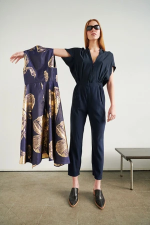 Model in a dark jumpsuit holding up a patterned dress against a plain wall in a campaign by Zero + Maria Cornejo