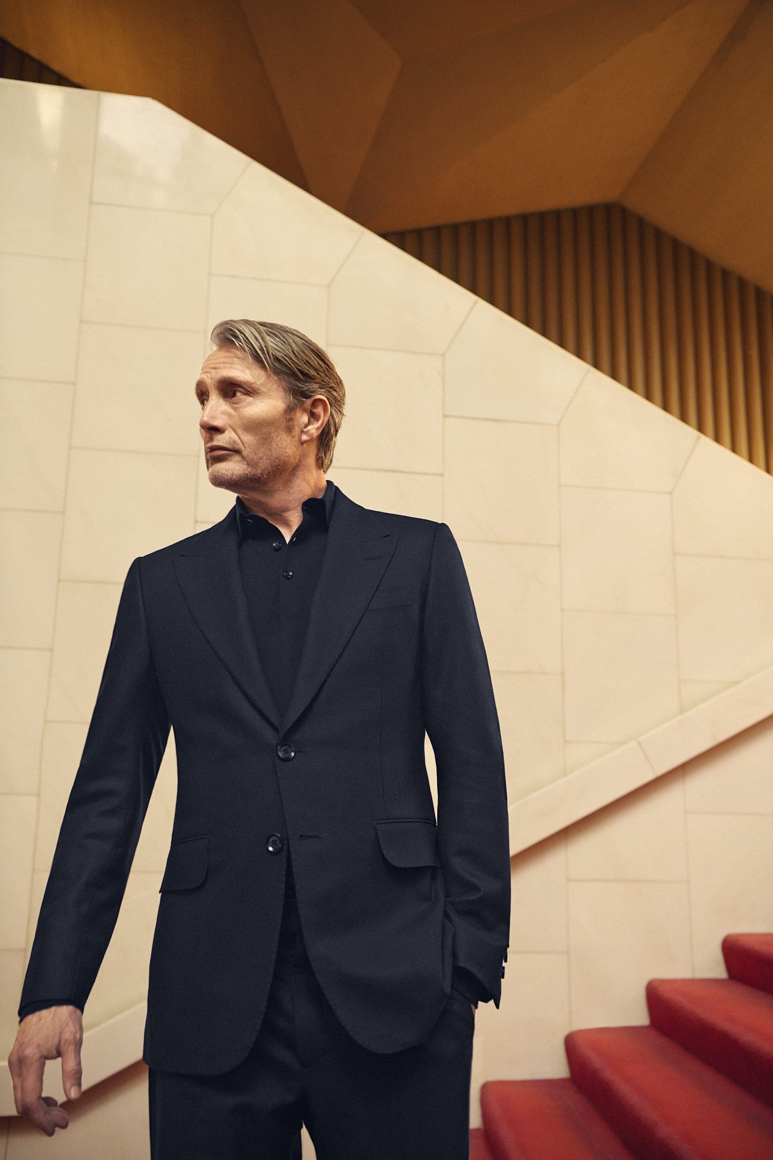 Mad Mikkelsen wearing a dark suit and shirt standing indoors near red stairs against a geometric architectural backdrop.