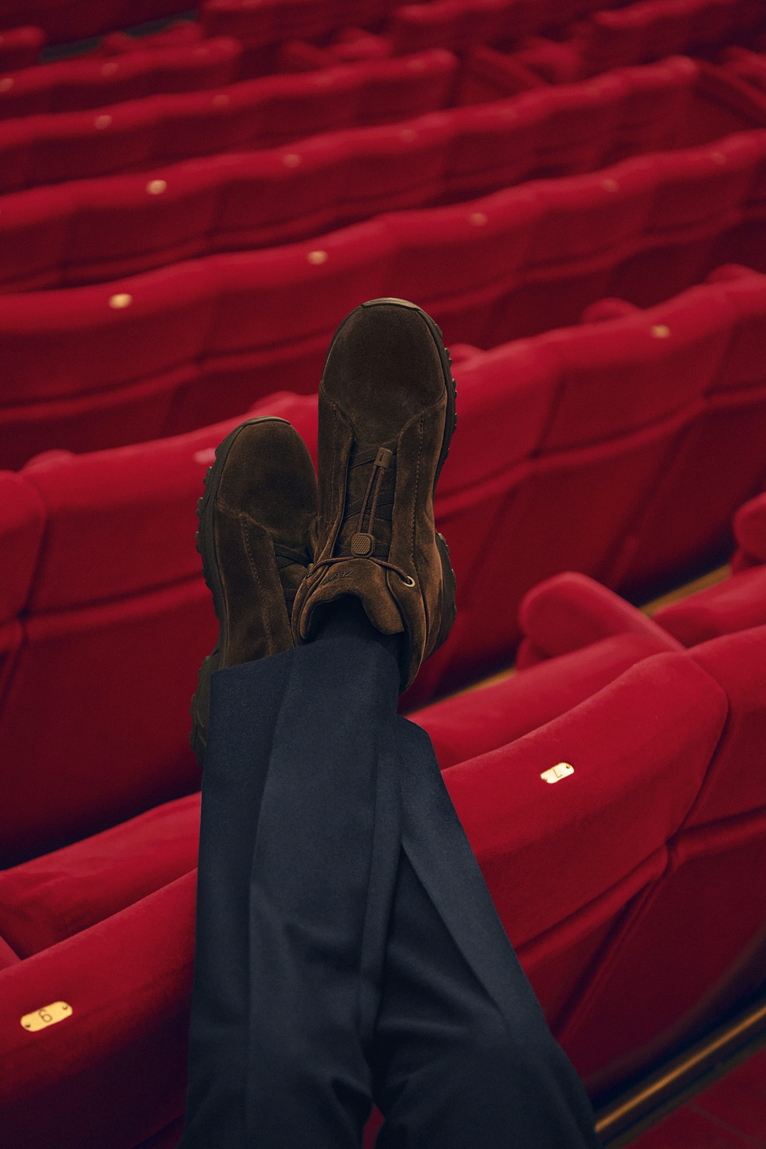 Feet up in brown suede shoes against rows of empty red theatre seats in an auditorium.