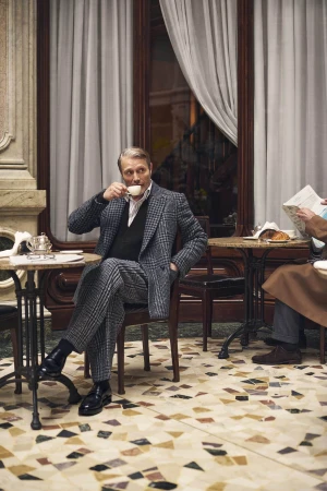 Mads Mikkelsen seated at a cafe table wearing a patterned grey tweed suit and drinking from a small cup.