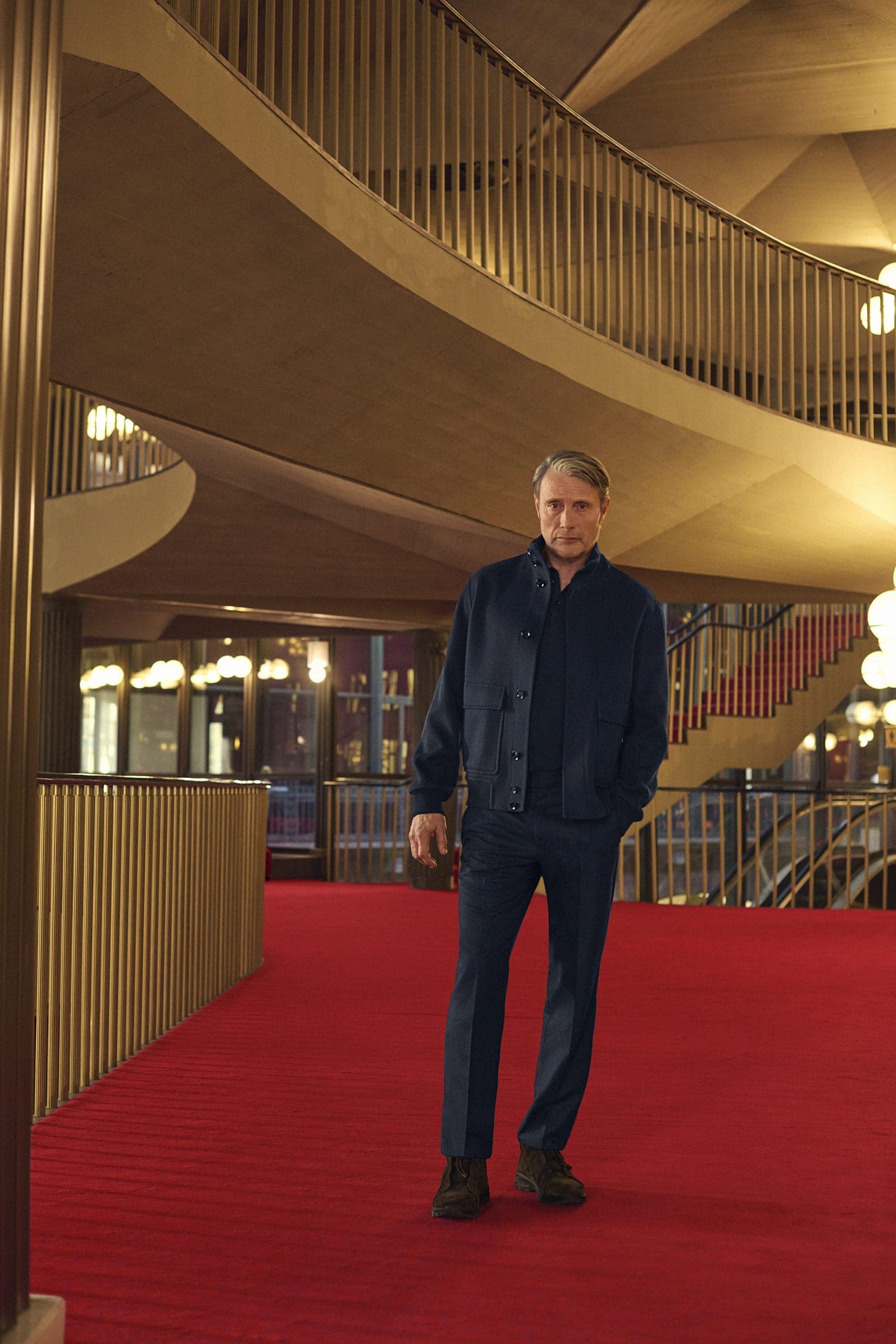Actor Mads Mikkelsen standing on a bright red carpet inside a grand modern architectural interior for Zegna Fall 2025 campaign.