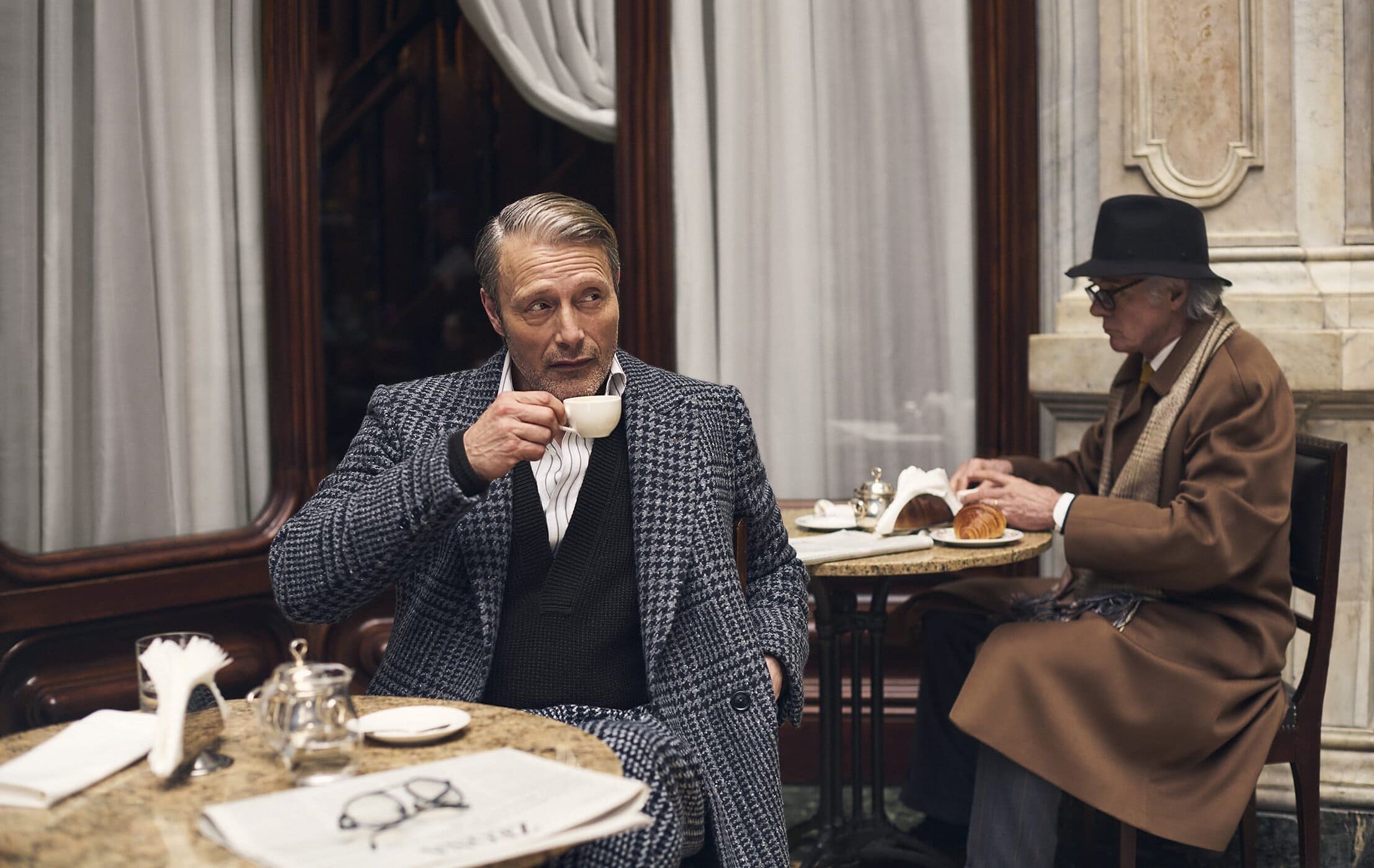 Mads Mikkelsen drinking from a small white cup in a houndstooth coat at a cafe table for Zegna Fall 2025 campaign.