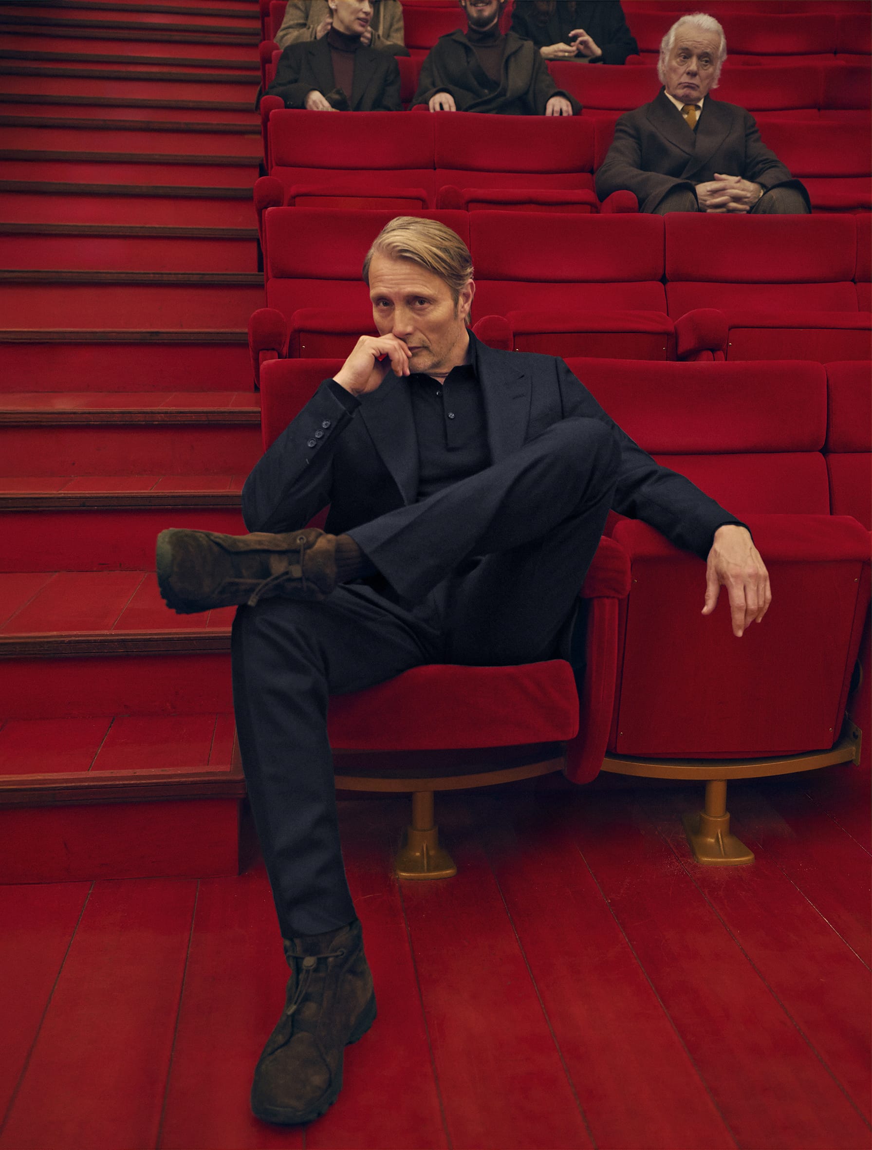 Mads Mikkelsen seated in a red theatre chair wearing a dark suit for the Zegna Fall 2025 campaign