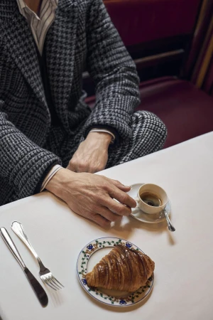 Mads Mikkelsen seated at a table wearing a houndstooth suit, reaching for a small espresso cup beside a croissant.