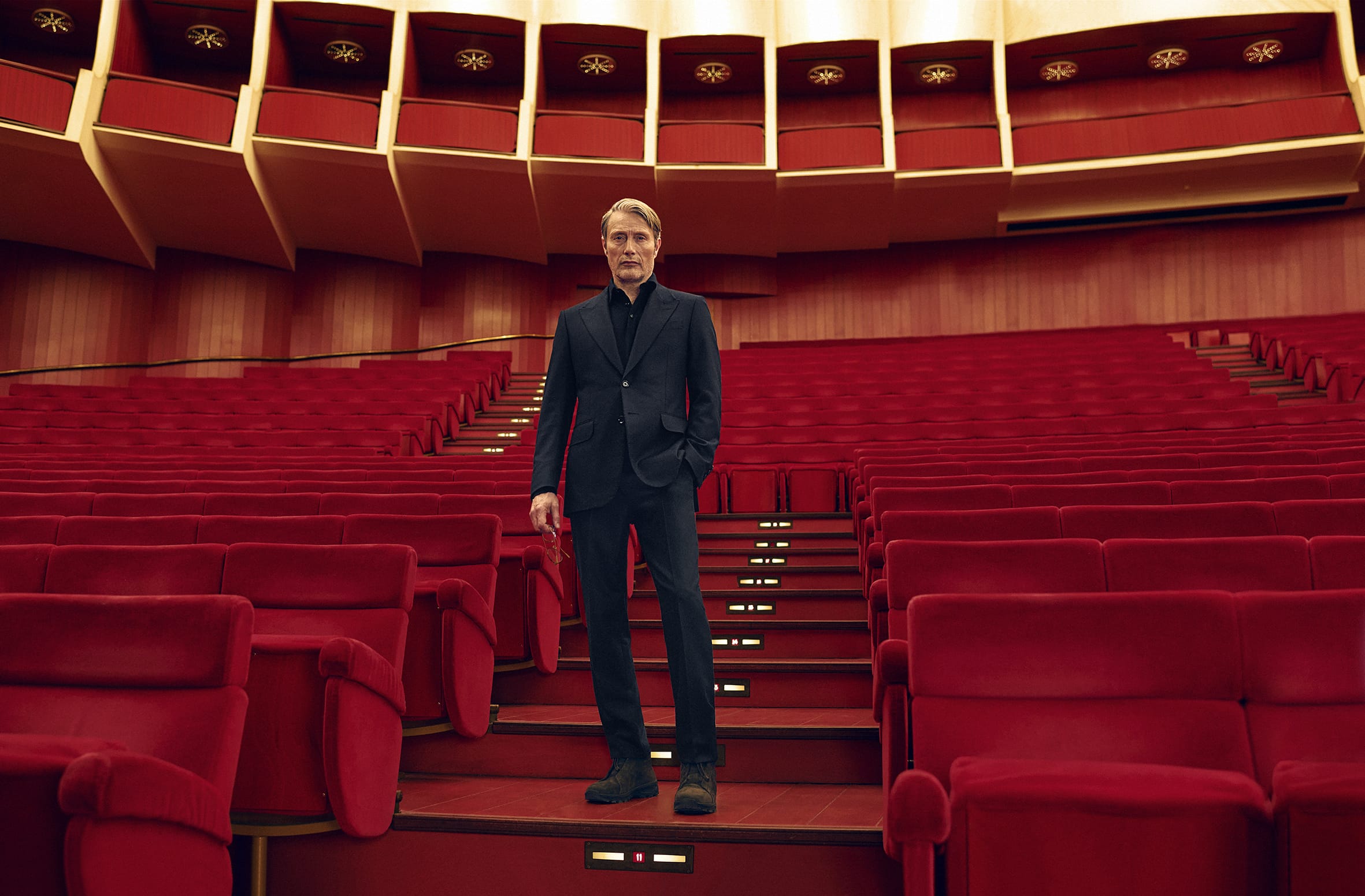 Mads Mikkelsen standing on red tiered seating inside the Teatro Regio di Torino wearing a dark suit for Zegna Fall 2025 ad campaign.