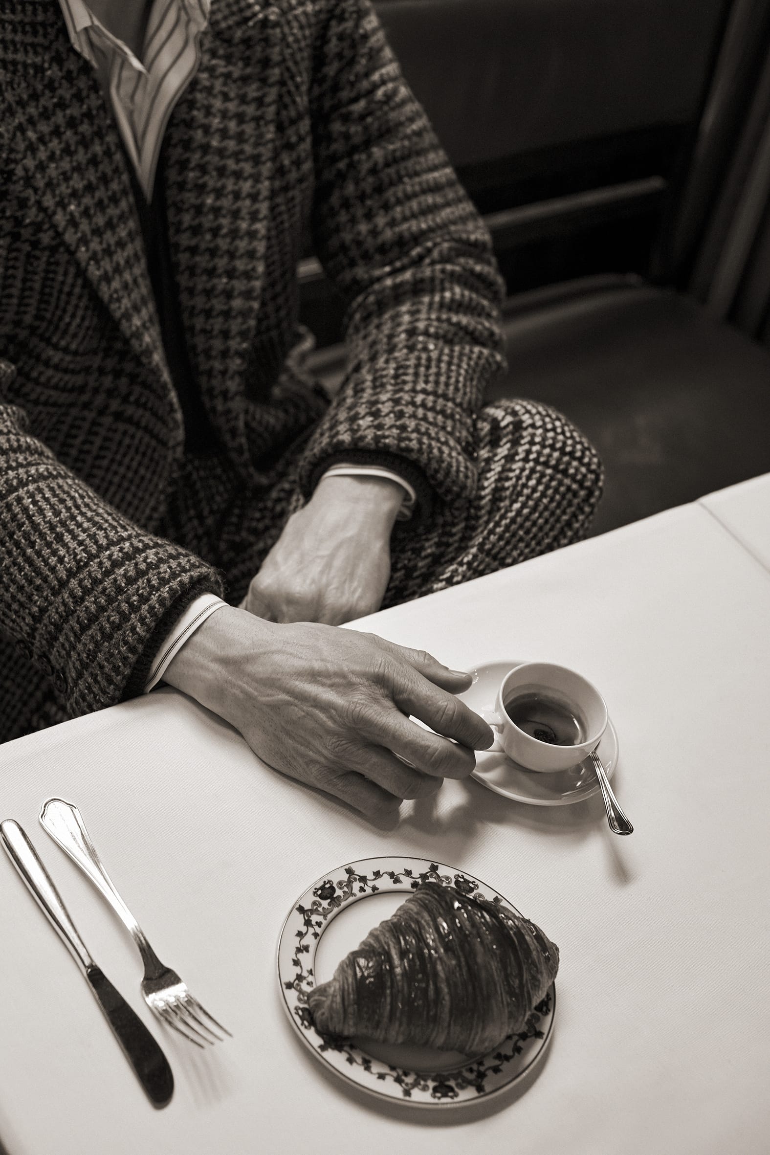 Mads Mikkelsen wearing a houndstooth suit sitting at a café table with a cup of coffee and a croissant