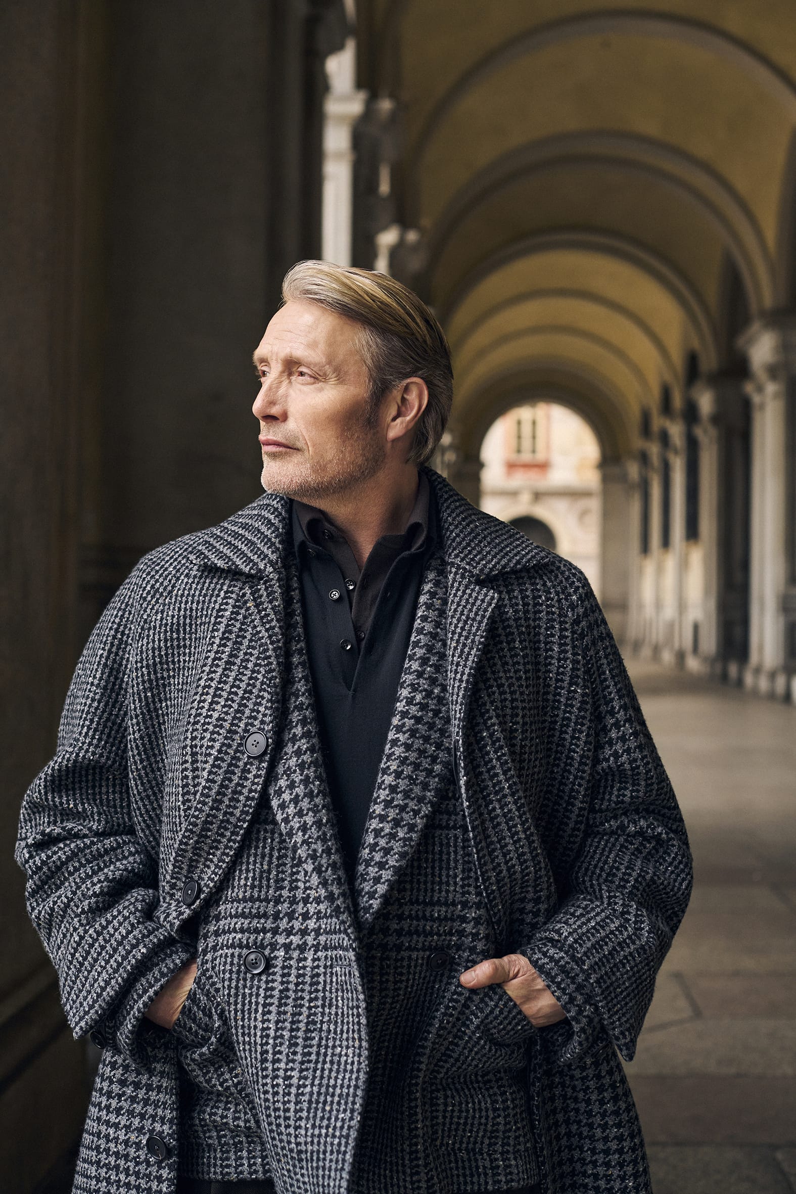 Mads Mikkelsen wearing a grey houndstooth coat and dark shirt, standing in an arched colonnade.